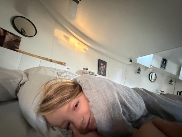A child with blonde hair peeks out from under a grey blanket, smiling softly while resting in bed